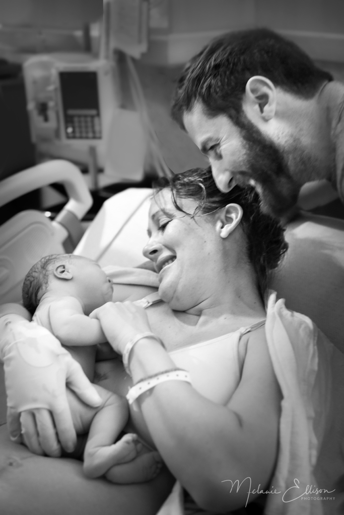 Woman in hospital bed holding newborn with father standing over mother and looking at baby.